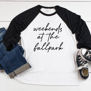May include: A white and black baseball tee shirt with the text "weekends at the ballpark" in black script. The shirt is laying on a white wooden surface with a pair of black and white sneakers and blue jeans.