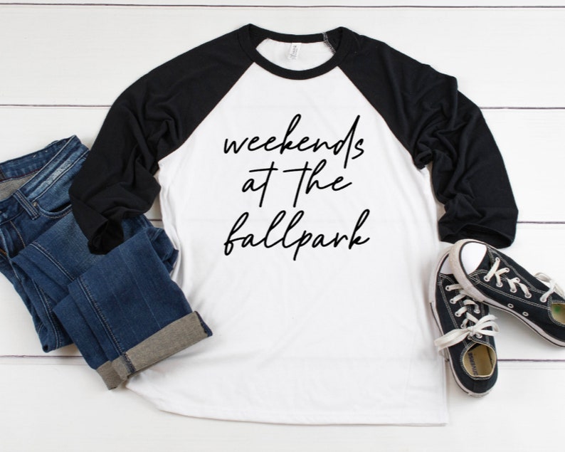 May include: A white and black baseball tee shirt with the text "weekends at the ballpark" in black script. The shirt is laying on a white wooden surface with a pair of black and white sneakers and blue jeans.