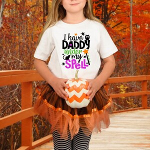 May include: A child wearing a white t-shirt with the text "I have Daddy under my spell" and a brown tulle skirt holds an orange and white pumpkin.