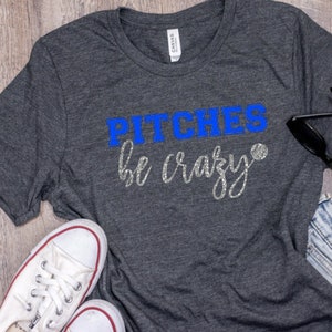 Puede incluir: Una camiseta gris con el texto "PITCHEs be crazy" en azul y purpurina plateada. Se incluye una pelota de béisbol en el texto.