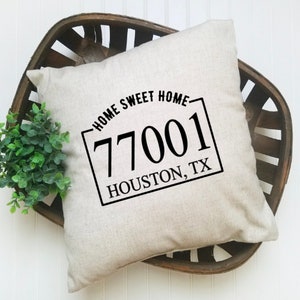 Könnte beinhalten: Ein beiger Dekokissen mit schwarzem Text, der "Home Sweet Home 77001 Houston, TX" lautet. Das Kissen liegt in einem Weidenkorb.