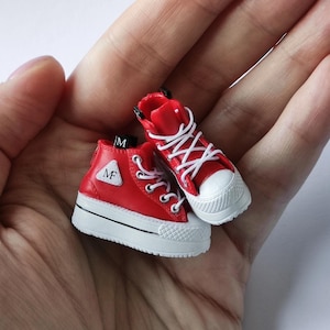 Peut inclure: Une paire de baskets rouges montantes avec des semelles et des lacets blancs. Les baskets ont un logo triangulaire noir avec les lettres "MF" à l'intérieur.
