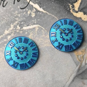 May include: Two circular buttons featuring a metallic blue finish and a clock face design. Each button displays raised Roman numerals from I to XII around the perimeter, with central clock hands and small circular markers for the hour positions.