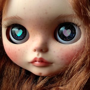 May include: A close-up of a doll's face with long, red hair. The doll has blue eyes with a pink heart design in each eye.