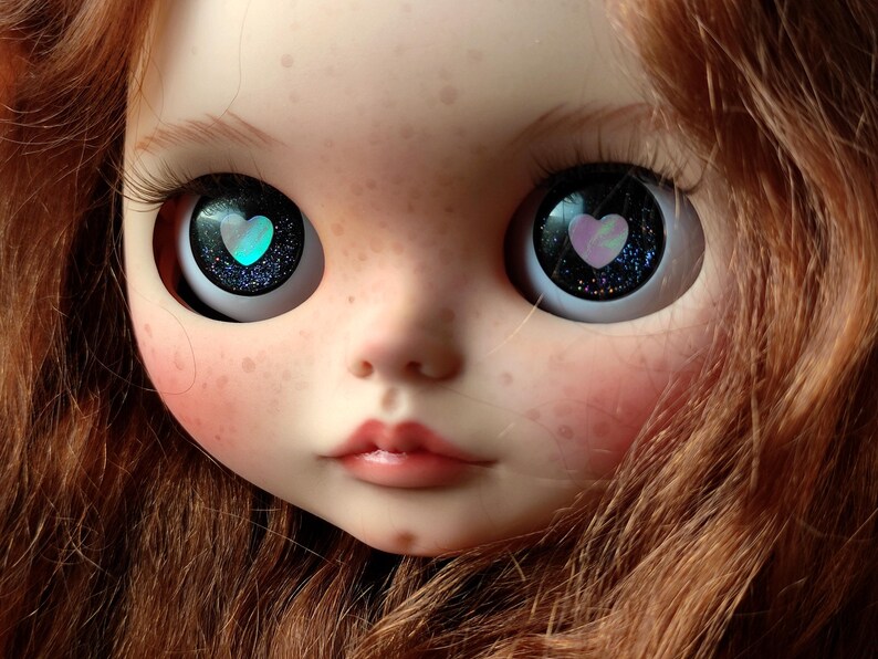May include: A close-up of a doll's face with long, red hair. The doll has blue eyes with a pink heart design in each eye.