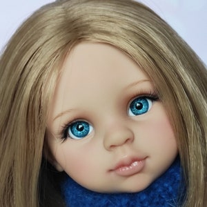 May include: A close-up of a doll's face with blonde hair and blue eyes. The doll is wearing a blue sweater.