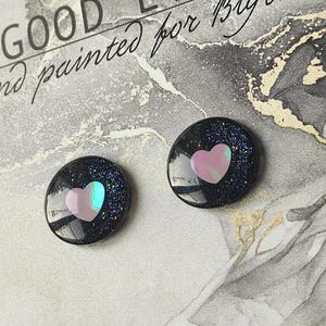 May include: A pair of black stud earrings with a pink heart design. The earrings are made of resin and have a glittery finish.