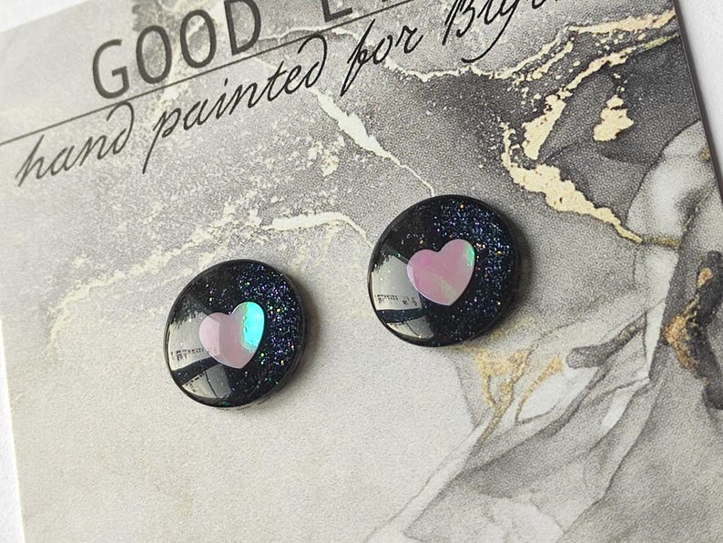 May include: A pair of black stud earrings with a pink heart design. The earrings are made of resin and have a glittery finish.