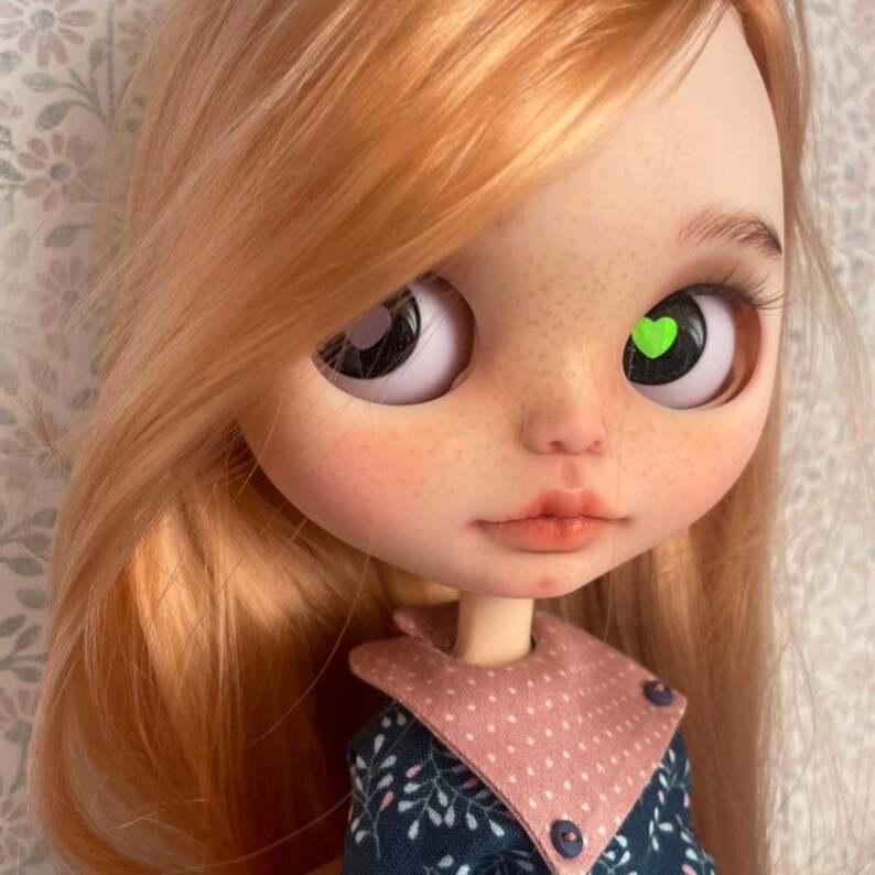 May include: A doll with long, straight, light brown hair. The doll has blue eyes with a green heart-shaped design on one eye. The doll is wearing a blue and white dress with a pink collar.