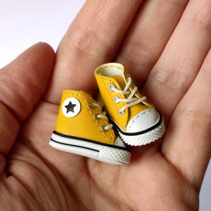 May include: Miniature yellow high-top sneakers with white soles and laces. Each shoe features a white star on a circular patch. The sneakers are held in a person's hand.