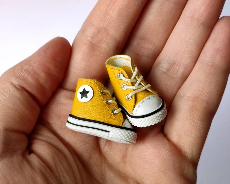 May include: Miniature yellow high-top sneakers with white soles and laces. Each shoe features a white star on a circular patch. The sneakers are held in a person's hand.