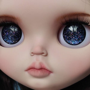 May include: Close-up of a doll's face with blue glitter eyes. The doll has a light pink complexion and a closed mouth.