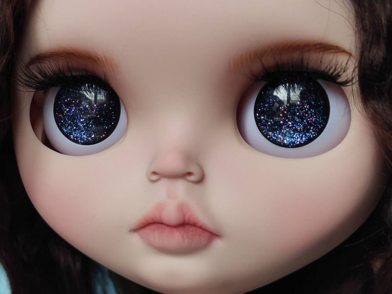 May include: Close-up of a doll's face with blue glitter eyes. The doll has a light pink complexion and a closed mouth.
