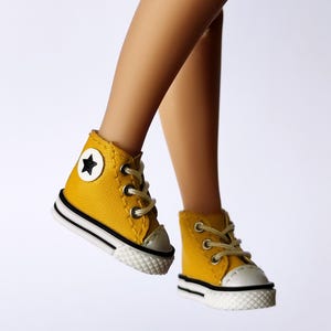 May include: A pair of miniature yellow high-top sneakers with white soles and black stripes. Each shoe features a white toe cap, tan laces, and a black star emblem on the outer side. The shoes are designed for a doll or similar figure.