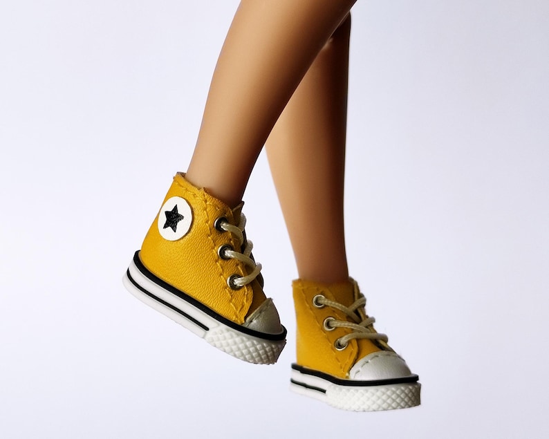 May include: A pair of miniature yellow high-top sneakers with white soles and black stripes. Each shoe features a white toe cap, tan laces, and a black star emblem on the outer side. The shoes are designed for a doll or similar figure.