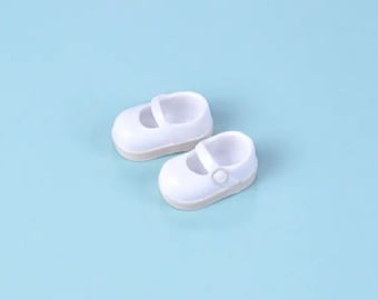 May include: A pair of white doll shoes with a buckle strap. The shoes are on a light blue background.