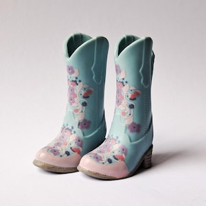 May include: A pair of light blue cowboy boots featuring a pale pink toe cap and a decorative floral pattern in shades of purple, white, and coral. The boots have a classic pull-on silhouette with a mid-calf height and a small, stacked block heel.