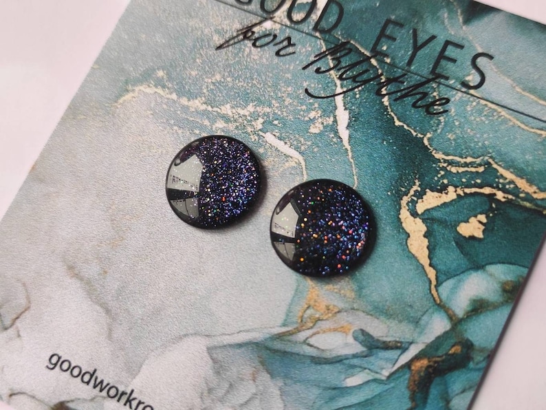 May include: A pair of black stud earrings with a glittery finish. The earrings are on a blue and gold background with the text "Good Eyes for the"