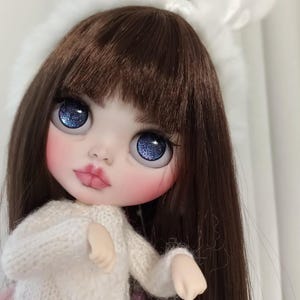 May include: A close-up of a doll with long brown hair, wearing a white knitted sweater. The doll has blue eyes with glitter and pink blush on her cheeks.