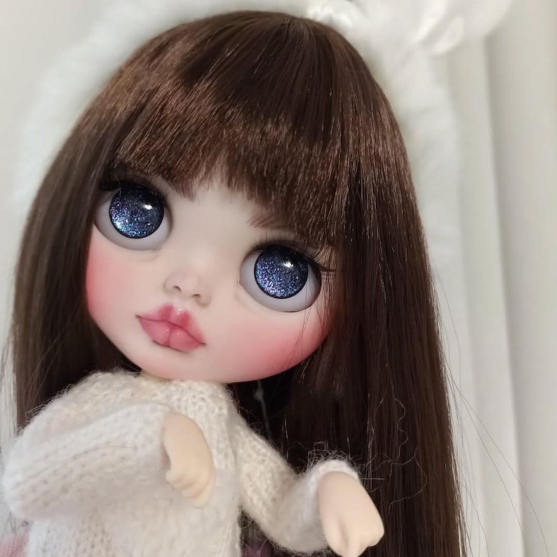 May include: A close-up of a doll with long brown hair, wearing a white knitted sweater. The doll has blue eyes with glitter and pink blush on her cheeks.