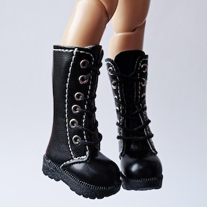 K&ouml;nnte beinhalten: Ein Paar schwarze Puppenstiefel mit wei&szlig;en N&auml;hten und silbernen &Ouml;sen. Die Stiefel sind mit schwarzen Schn&uuml;rsenkeln geschn&uuml;rt und haben eine strukturierte Sohle. Die Stiefel sind etwa 5 cm hoch.