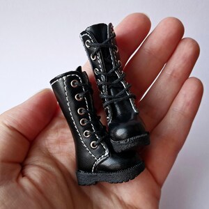 K&ouml;nnte beinhalten: Ein Paar Miniatur-Stiefel in Schwarz mit wei&szlig;en N&auml;hten und Schn&uuml;rsenkeln. Die Stiefel haben ein klassisches Combat-Boot-Design mit mehreren &Ouml;sen f&uuml;r die Schn&uuml;rsenkel und einer strukturierten Sohle. Die Stiefel sind wahrscheinlich f&uuml;r eine Puppe oder eine andere kleine Figur.