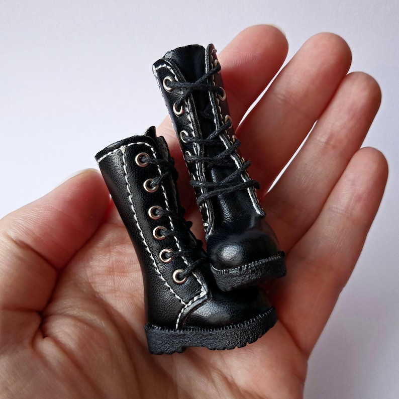 K&ouml;nnte beinhalten: Ein Paar Miniatur-Stiefel in Schwarz mit wei&szlig;en N&auml;hten und Schn&uuml;rsenkeln. Die Stiefel haben ein klassisches Combat-Boot-Design mit mehreren &Ouml;sen f&uuml;r die Schn&uuml;rsenkel und einer strukturierten Sohle. Die Stiefel sind wahrscheinlich f&uuml;r eine Puppe oder eine andere kleine Figur.