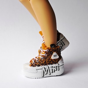 May include: A high-top trainer with a brown and black leopard print pattern, white laces, and a thick white platform sole. The side of the sole features the word MINI in black lettering, and a small white triangular patch displays the letter M.