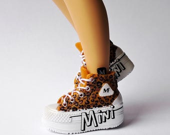 Può includere: Una scarpa alta con stampa leopardata marrone e nera, lacci bianchi e una spessa suola platform bianca. Il lato della suola presenta la parola MINI in lettere nere e una piccola toppa triangolare bianca mostra la lettera M.