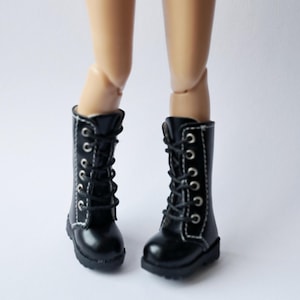 K&ouml;nnte beinhalten: Ein Paar schwarze Puppenstiefel mit wei&szlig;en N&auml;hten und silbernen &Ouml;sen. Die Stiefel sind geschn&uuml;rt und haben einen kleinen Absatz. Die Stiefel sind an den Beinen einer Puppe zu sehen.