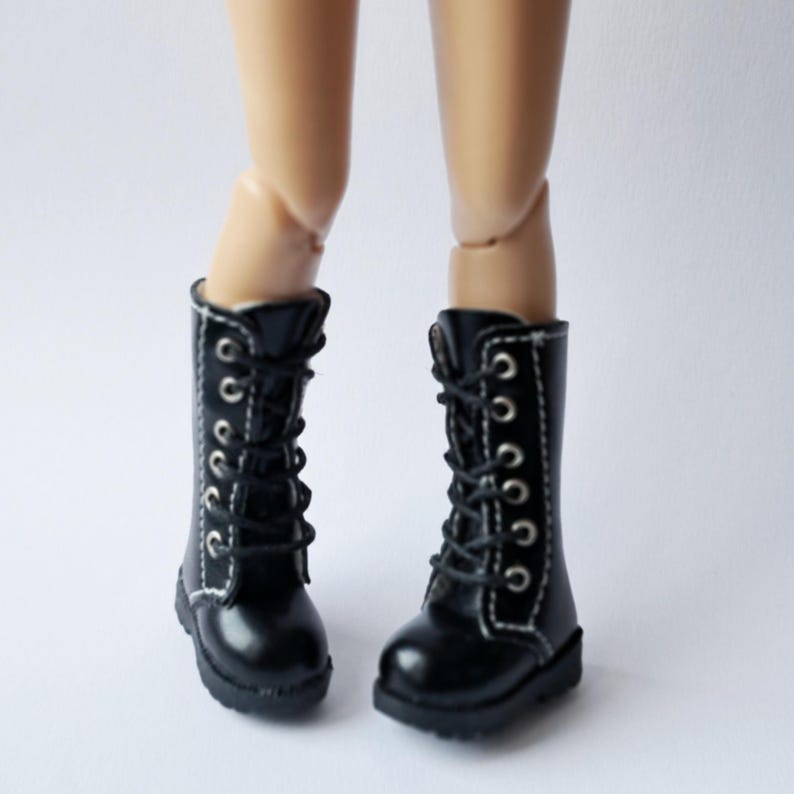 K&ouml;nnte beinhalten: Ein Paar schwarze Puppenstiefel mit wei&szlig;en N&auml;hten und silbernen &Ouml;sen. Die Stiefel sind geschn&uuml;rt und haben einen kleinen Absatz. Die Stiefel sind an den Beinen einer Puppe zu sehen.