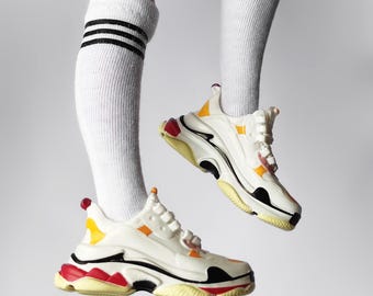 Può includere: Sneakers bianche con dettagli neri, rossi e gialli. Le sneakers hanno una suola spessa e robusta e sono indossate con calzini bianchi a righe nere.