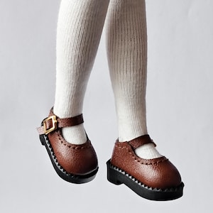 May include: A pair of brown leather doll shoes with black soles and a gold buckle. The shoes have a strap across the top and a decorative stitch around the edge.