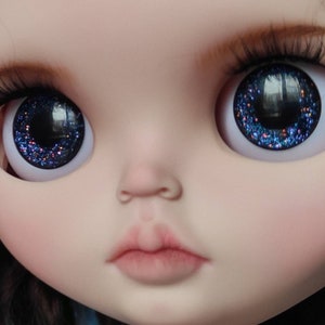 May include: Close-up of a doll's face with blue glitter eyes. The doll has a light pink complexion and a closed mouth.