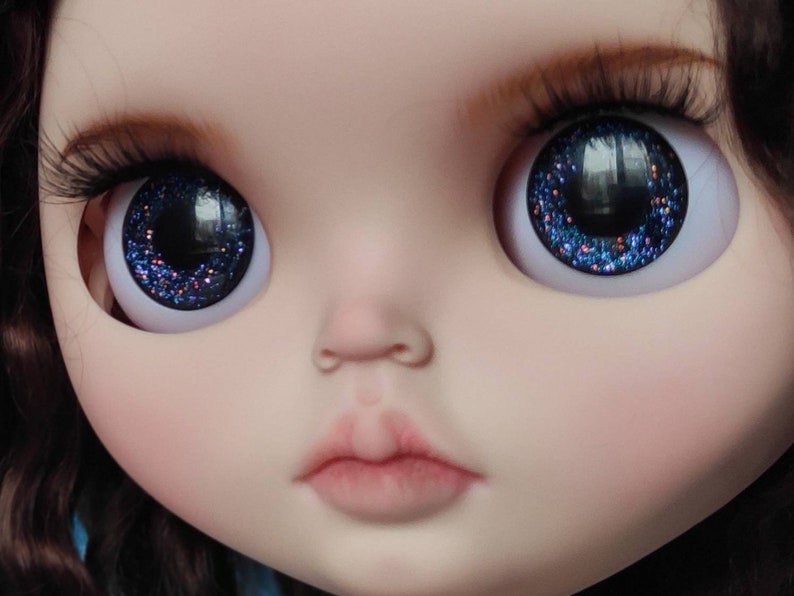 May include: Close-up of a doll's face with blue glitter eyes. The doll has a light pink complexion and a closed mouth.