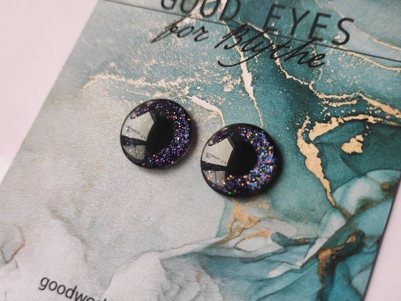 May include: Two black plastic eyes with iridescent glitter. The eyes are round and have a black center. The eyes are on a blue and white background with the text "GOOD EYES for Blythe".