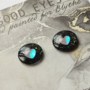 May include: Two black resin doll eyes with a pink and blue heart design. The eyes have a glittery finish and are hand painted.