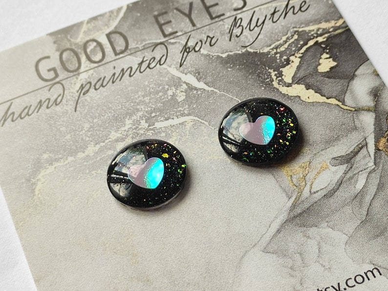 May include: Two black resin doll eyes with a pink and blue heart design. The eyes have a glittery finish and are hand painted.
