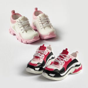Può includere: Due paia di scarpe da ginnastica. Un paio è bianco con accenti rosa e l'altro paio è nero, bianco e rosso.