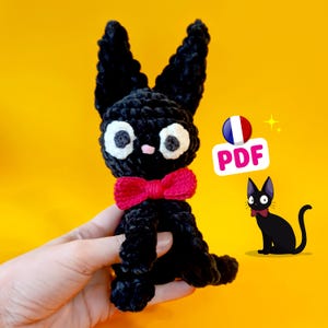May include: A handmade black crochet cat with large white eyes, a pink nose, and a red bow tie. The cat has large, upright ears and is sitting. A cartoon illustration of a black cat with a red bow tie is next to the crochet cat. The image has a yellow background.