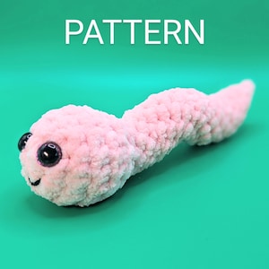May include: A pink crocheted worm with black eyes and a smiling mouth. The worm is lying on a green surface. PATTERN is written in white letters above the worm.