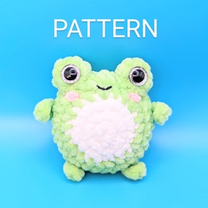 May include: A green crocheted frog plush toy with white fur on its belly and large black eyes. The toy is sitting on a blue background.