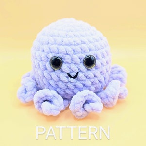 May include: A crocheted purple octopus with black eyes and a smiling mouth. The octopus is sitting on a light yellow background.