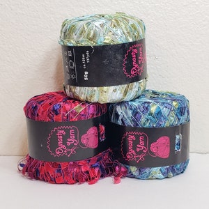 Könnte beinhalten: Drei Knäuel Garn in verschiedenen Farben. Die Knäuel sind mit "Dynasty Yarn" beschriftet und haben ein pinkfarbenes Herzlogo. Das Garn ist eine Mischung aus Grün, Blau und Rot.