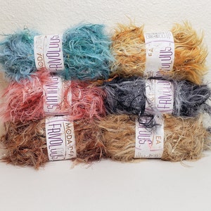 Pode incluir: Seis novelos de fio em várias cores, incluindo turquesa, rosa, marrom, bege e cinza. Cada novelo é etiquetado com "Fabulous" com o nome da marca "Moda-Crea" em texto roxo.
