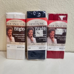 Op de afbeelding: Drie verpakkingen Wrights biaisband in wit, marineblauw en baksteenrood. De verpakkingen tonen de productnaam, de lengte in yards en meters en de productcode. De witte verpakking is gemarkeerd met "Satin Bias Tape".