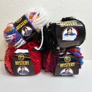 May include: Four skeins of Lion Brand Yarn Mystery yarn. Each skein is a different color combination: red and orange, purple and red, black and white, and rainbow. Each skein has a label with a woman wearing a scarf and the words "Lion Brand Yarn" and "Mystery".