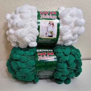 Könnte beinhalten: Zwei Knäuel Bernat Puff Ball Holidays Garn. Eines ist weiß, das andere grün. Das Garn hat ein strukturiertes, puffiges Aussehen. Die Etiketten tragen die Aufschrift "Puff Ball Holidays" und zeigen ein Kind mit einem Pullover.