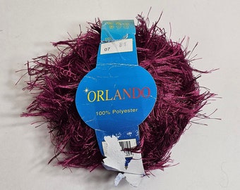 Può includere: Un gomitolo di filato color bordeaux dalla consistenza lanuginosa. Un'etichetta blu con la scritta "ORLANDO" e "100% Poliestere" è attaccata. L'etichetta riporta anche i numeri "07" e "81".