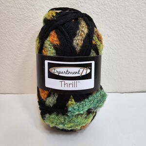 Puede incluir: Un ovillo de hilo Thrill de Department 71. El hilo es una mezcla de colores negro, verde, naranja y beige. La etiqueta es negra con texto blanco. El hilo está envuelto en una banda negra con el nombre de la marca y del producto.